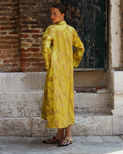Woman in a yellow coat standing against a brick wall.