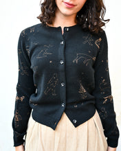 Load image into Gallery viewer, A woman wearing a dark charcoal knit cardigan with gold line-art illustrations of astronauts, planets, and stars, paired with a beige pleated skirt, against a plain white background.
